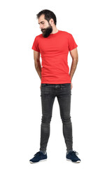 Serious bearded hipster in red t-shirt with hands in his back pocket looking down. Full body length portrait isolated over white studio background.