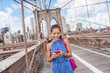 Happy Asian woman using phone texting walking on Brooklyn bridge, New York city for social media or blogging. Tourist doing summer travel in Manhattan, USA.