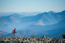 Hiker Is Walking On The Rocky Mountain On Backpacking Trip. Man Is Wearing Red Jacket And Backpack On. Beautiful Mountains On Background. Ecotourism And Healthy Lifestyle Concept. Copy Space.