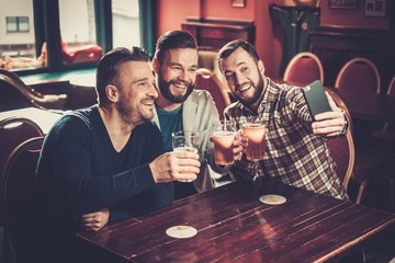 Wall Mural - Cheerful old friends having fun taking selfie and drinking draft beer in pub.