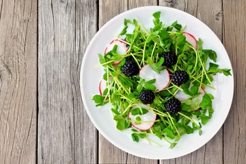 Healthy salad with pea shoots, radishes and blackberries on white plate against a rustic wood background