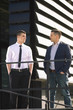 © bokan - Two young businessmen portrayed in front of the modern office building. They are standing with their hands in pockets and looking at each other.