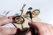 Miniature Of Wooden Bicycle On White Background. Handcrafting Process, Craftsman's Hand Holding The Tool. Macro Shot.