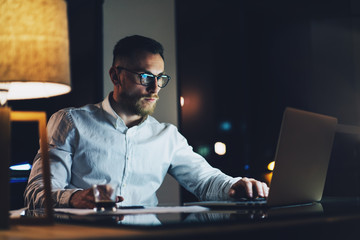 Wall Mural - Bearded young businessman wearing white shirt working on modern loft office at night. Man using contemporary notebook texting message, blurred background. Horizontal, film effect. 