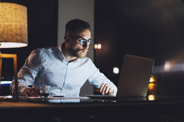 Wall Mural - Bearded businessman wearing white shirt working on modern loft office at night. Man using contemporary notebook texting message, blurred background. Horizontal, film effect, bokeh
