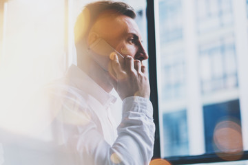 Portrait bearded adult businessman working on modern loft office. Man wearing white shirt and using contemporary smartphone. Panoramic windows background. Horizontal, film effect, bokeh