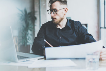 Photo bearded adult businessman working on modern urban cafe. Man wearing black shirt and looking laptop, holding papers pencil. Horizontal, film effect, bokeh. 