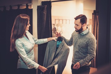 Wall Mural - Confident handsome man with beard choosing a jacket in a suit shop.