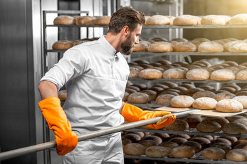 Handsome baker in uniform with orange working gloves putting with shovel from the oven bread loafs on the shelves at the manufacturing