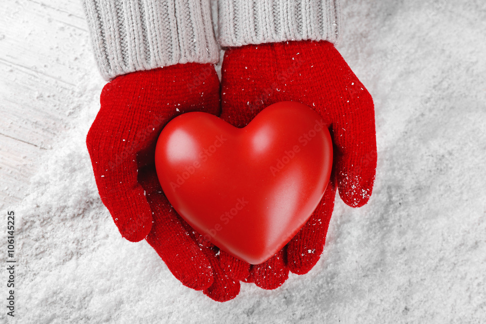 Hands in warm red gloves holding heart on snowy background