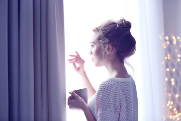 Blonde beautiful  girl looking in the window and holding a cup of coffee or tea in her hands