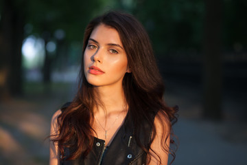 Wall Mural - Closeup portrait of happy young beautiful brunette woman in black leather jacket posing on sunset outdoors  with blurry foliage background