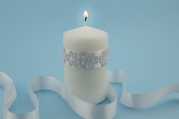 Photo of single white lit candle isolated with white satin ribbon blue background