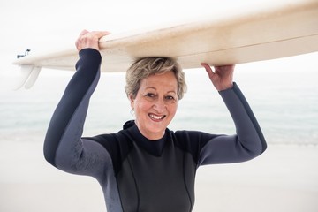 Wall Mural - Portrait of senior woman in wetsuit carrying surfboard over head