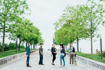 Wall Mural - Group of young multiethnic friends standing outdoor ina city park, turning looking in camera, holding bike and skate, smiling - happiness, friendship concept - copy space on top