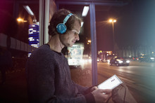 Germany, Munich, Man With Headphones Sitting At Bus Stop Using Digital Tablet At Night