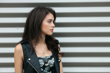 Wall Mural - Closeup portrait of young beautiful brunette woman in black leather jacket posing outdoors against urban style background of metal strips