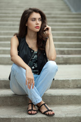 Wall Mural - Young thoughtful fashionable brunette woman in black leather jacket posing sitting on concrete stairway