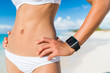 © Maridav - Fit bikini woman wearing wearable tech smartwatch. Closeup of slim abs stomach and beachwear on of female person relaxing on beach with activity tracker wrist band on summer holidays.