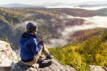 Wall Mural - tourist young woman is sitting with a mug of tea on a cliff overlooking the autumn mountains with fog. The concept of a happy holiday in the nature, Hiking