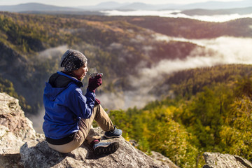 tourist young woman is sitting with a mug of tea on a cliff overlooking the autumn mountains with fo