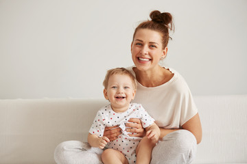 charming brunette woman playing with her baby while sitting on a bed in her apartment at the weekend