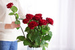 Woman putting red roses into vase indoors