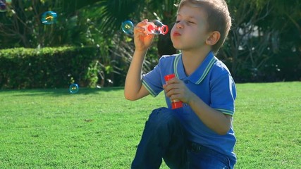 Wall Mural - Little boy blowing soap bubbles on fresh green grass field, having fun in the park in sunny day, slow motion, happy carefree childhood. Full HD Video 1920x1080