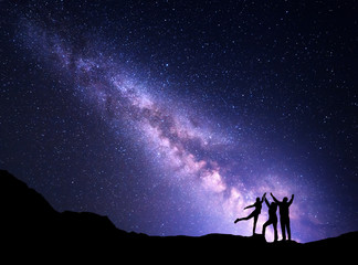Wall Mural - Landscape with purple Milky Way. Night starry sky with silhouette of a happy family with raised-up arms on the mountain. Beautiful Universe. Space background
