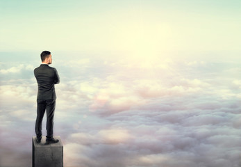 Young man in business suit, standing on concrete column above clouds.