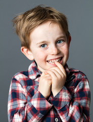 portrait of a shy red hair 5-year old boy smiling and biting his teeth for childhood and wellbeing, grey background studio