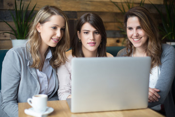 Wall Mural - Three young women sitting at cafe drinking coffee and looking at laptop
