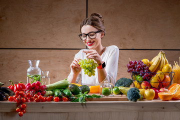 Wall Mural - Young and cute woman eating grapes at the table full of fruits and vegetables in the wooden interior. Healthy food concept. Beauty and wellbeing