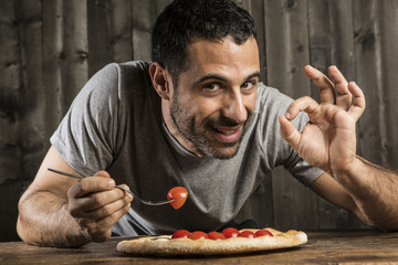 Ragazzo con i capelli scuri con mangia  pizza con i pomodorini e fà il segno di ok - sfondo legno scuro e  tavolo di legno chiaro