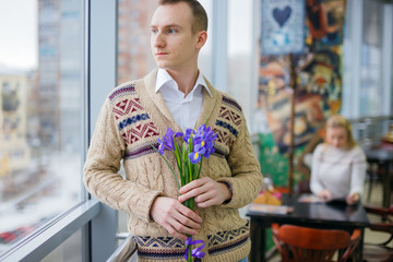 sad in love guy with holding flowers waiting for someone. man waiting for the boyfriend or girlfriend on a date. behind the man lonely girl sitting at a table in a cafe.