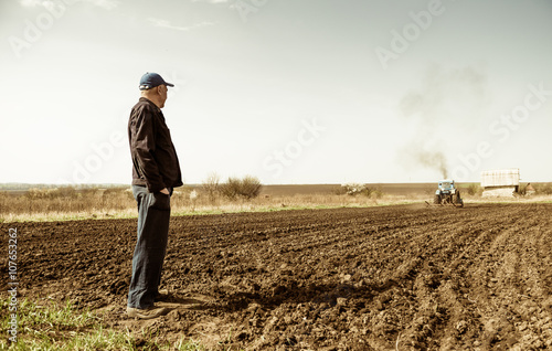 фотография farmer looking at tractor plowing ground at spring season