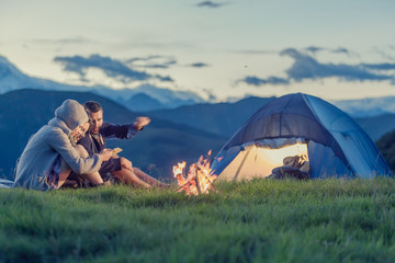 three friends camping with fire on mountain at sunset