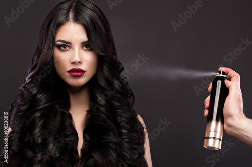 Zdjęcie XXL Piękny model brunetka: loki, klasyczny makijaż i czerwone usta z butelką produktów do włosów. Piękna twarz.