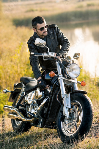 Fotomural Hombre brutal joven en una chaqueta negro y gafas de pie cerca de una motociclet