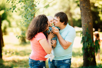 Wall Mural - Young mother and father kissing little son in park. Happy family with kids having picnic outdoors. Parents with children relax in a sunny summer garden. 