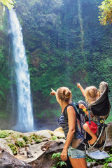 In waterfall pool young happy mother hold little traveller - baby girl in carrying backpack on back. Travel adventure, hiking activity with child. Lifestyle on family summer vacation and weekend tour