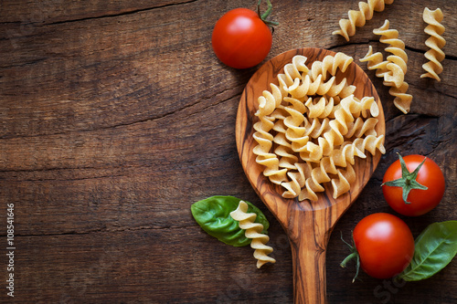 Fotografie, Obraz Italian cuisine concept - Wholemeal farfalle pasta in wooden spoon with fresh basil and tomatoes od dark rustic wooden background