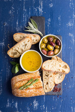 Mediterranean Snacks Set. Olives, Oil, Herbs And Sliced Ciabatta Bread On Black Slate Stone Board Over Painted Dark Blue Background