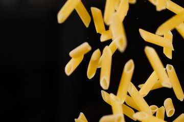 Falling penne pasta. Flying yellow raw macaroni over black background.