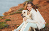 Fototapeta Las - A beautiful young woman,brunette with blue eyes and curly hair,dressed in a white jacket and light trousers, spends time on a rocky beach with his faithful friend, the dog breed white Labrador