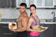 Fit Couple Looking Angry In The Kitchen; Animal Versus Plant Proteins: One Plate With Beef, Eggs, Salmon, Cheese And Chicken Grill And Another With Nuts, Mushrooms, Broccoli, Lentil, Hummus And Quinoa