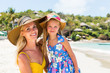 Close-up of a beautiful mother woman in yellow dress and beach straw hat with her pretty blonde little daughter at tropic seaside. Both smile to camera. Happy family on the beach. Happy mothers day.