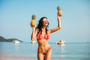 Wall Mural - Close up fashion summer portrait of stylish joyful happy sexy woman with perfect tanned fit body wearing trendy sunglasses, holding a pineapple, tropical fruit.