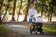 Young Mother Walking And Pushing A Stroller In The Park. Mother Strolling With Newborn. Beautiful Happy Mother With Pram Outdoors. Summer Walks In The Sunny Day. Girl With Carriage.