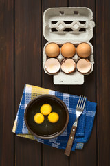 Wall Mural - Three raw eggs in rustic bowl with fork on the side and egg box with eggs and eggshells above, photographed on dark wood with natural light